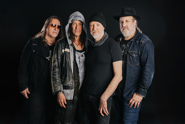 Four older men taking a group photo. They are wearing darker clothes and standing in front of a black backdrop.