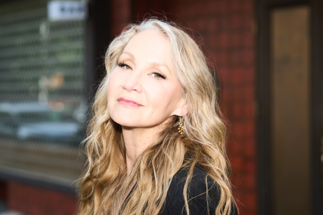 A woman with long, wavy blonde hair and light makeup stands outside in soft sunlight, wearing gold earrings and a black top, with a calm expression. The background features a red brick wall and a glass window.